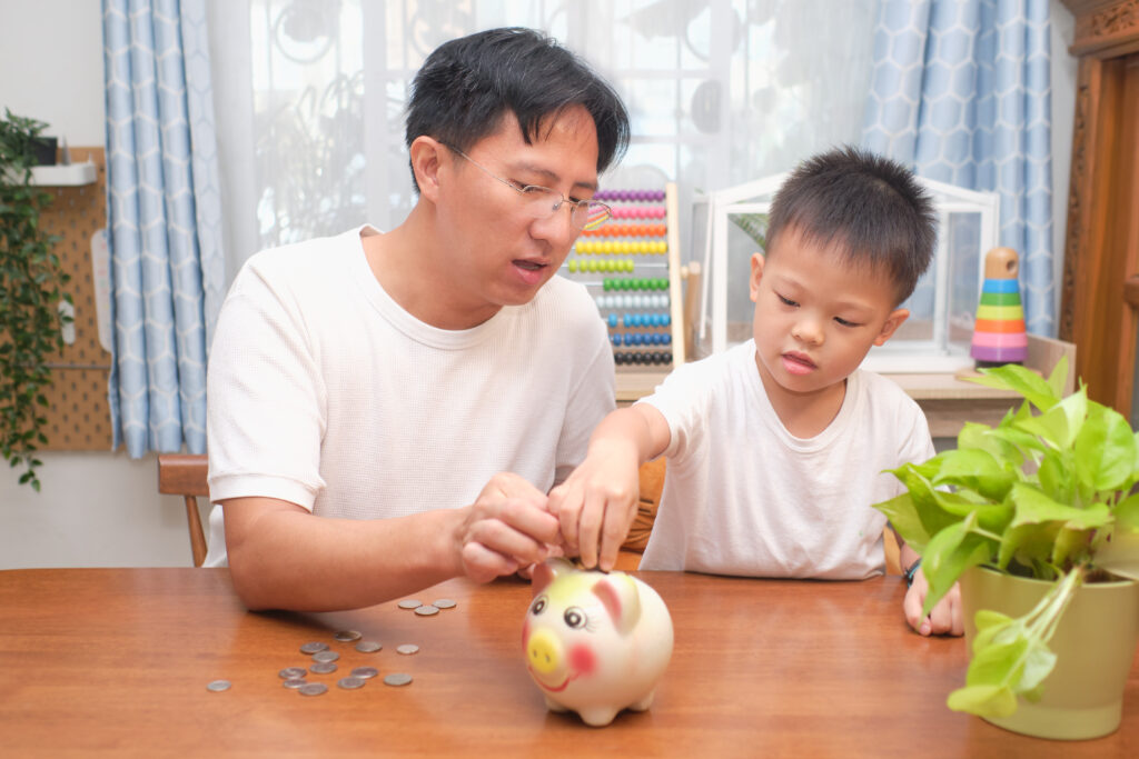 Father and son putting coins into piggy bank at home, family money savings concept. Dad teaching son on savings & financial planning, Little 5 years old kid saving money for future concept